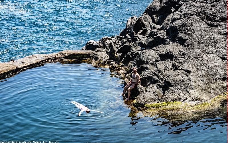 A diverse group of Pakistanis (men and women) engaged in discussion, some looking at smartphones or newspapers, possibly in a busy street market or chai dhaba setting
